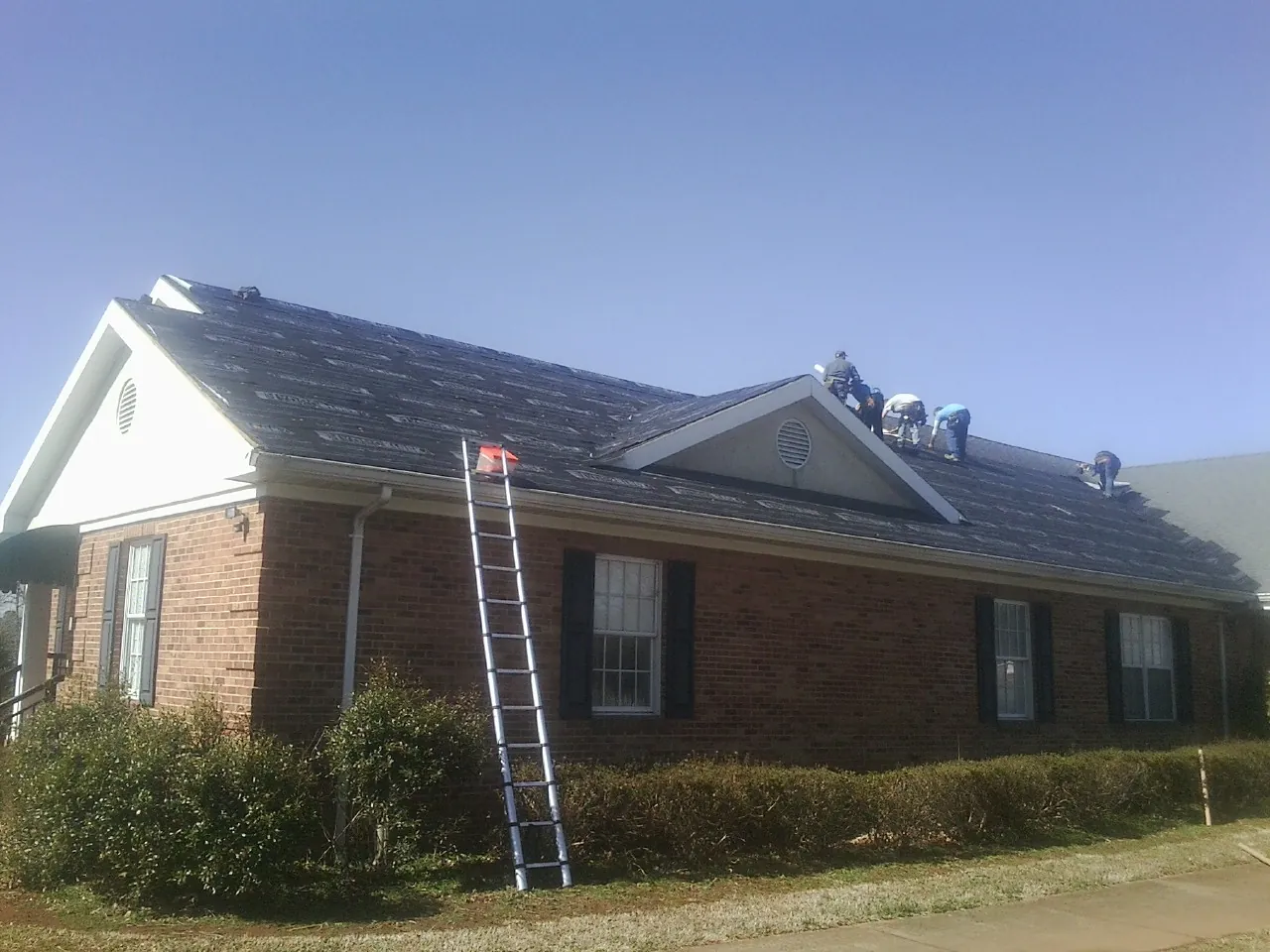 Crew tearing shingles off a brick ranch roof with a ladder set — Metal Roof Repair work by our crew in Grandin