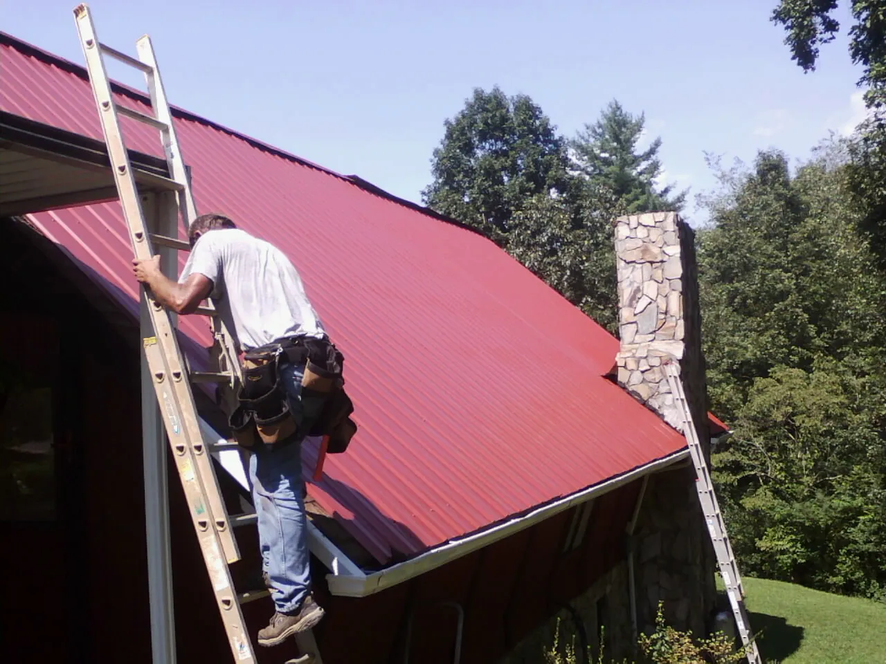 Roofer on a ladder beside a red metal roof and stone chimney — Soffit & Fascia Repair by Roofing Brothers in Grandin